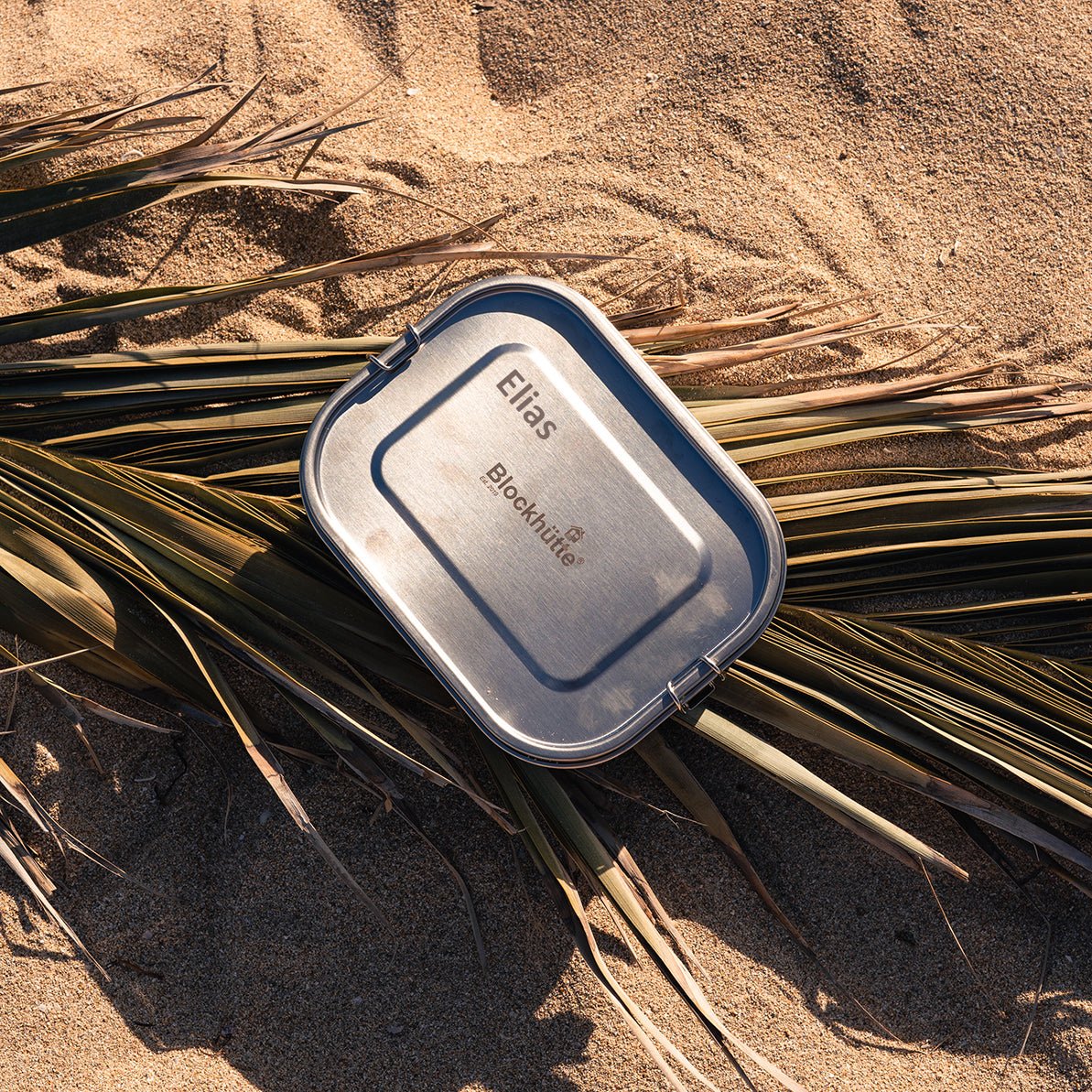 Boîte à repas en inox avec la gravure laser « Elias » sur le couvercle, dans le sable entre des palmes